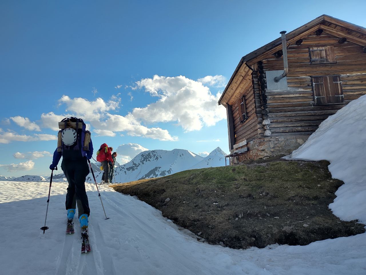 Friend and me skiing up to a hut in the Alps. Not a lot of snow left - we had to hike the first half in our ski boots. You can see a wooden hat, snow, mountains, and people from behind skinning up to the hat. They just arrived.