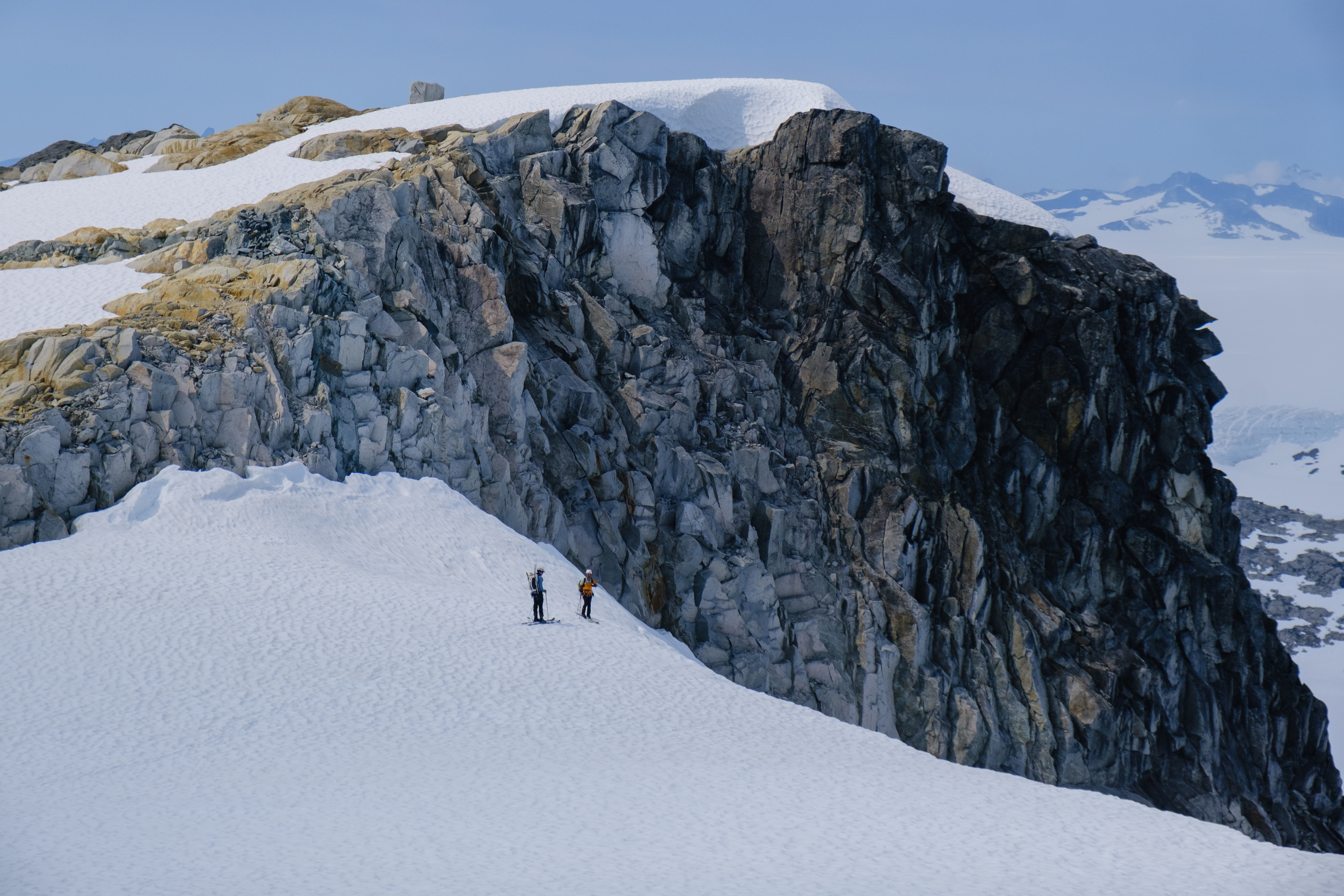 Two skiers looking deep down where a glacier drops many meters, all snow-covered. (Not an ice-fall). A rocky hill next to them lets you see how big everything is.