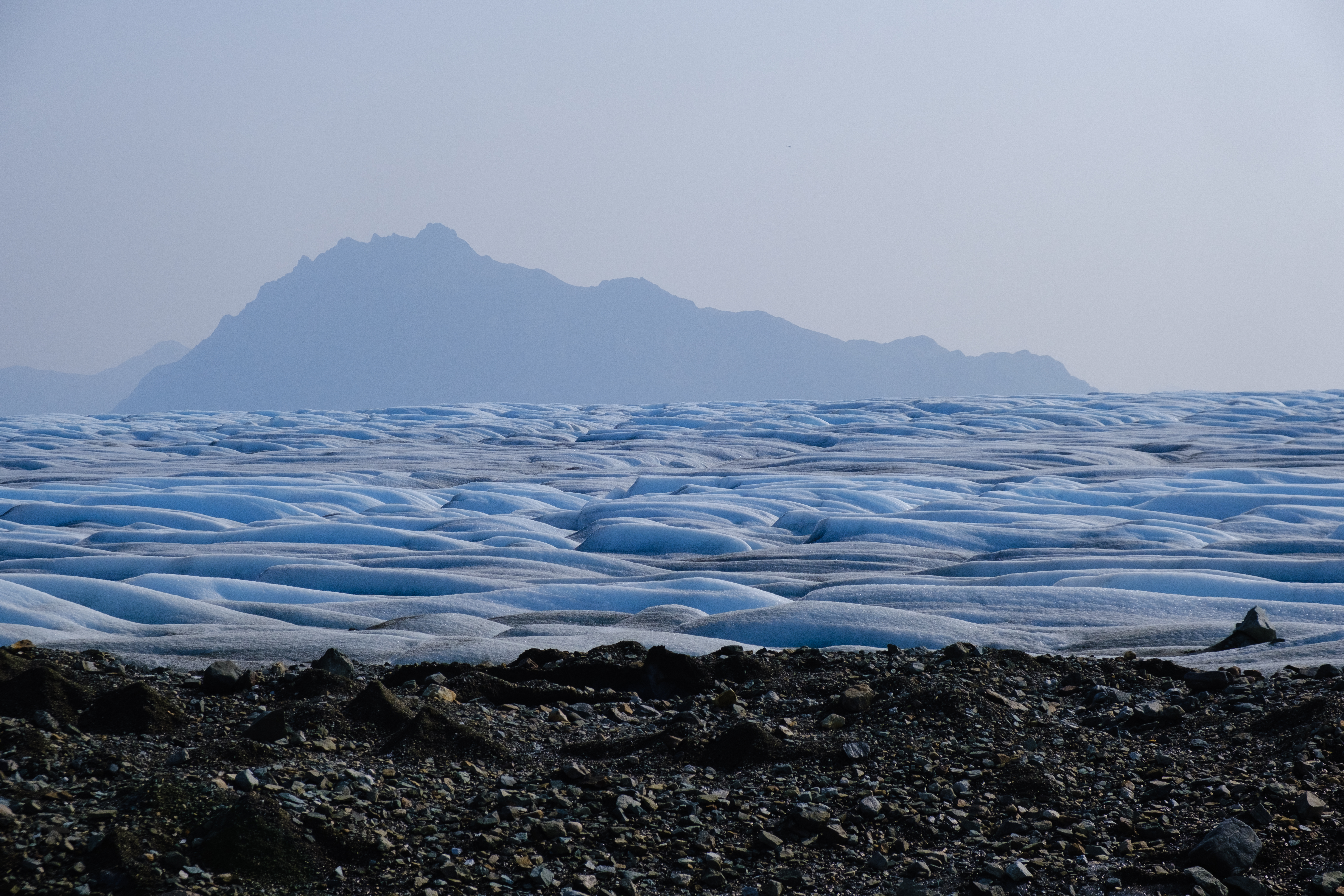 A bare glacier without snow, but the ice flows in soft waves. A mountain is in the haze in the background.